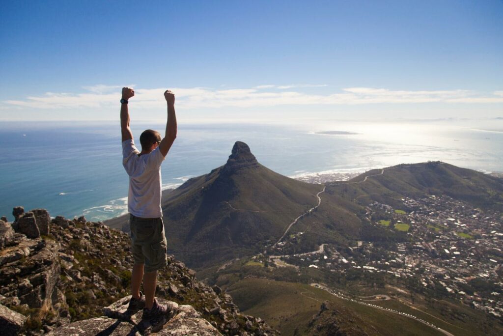 Man on top of Table Mountain with his arms in the air. Lions Head and part of Cape Town in the background